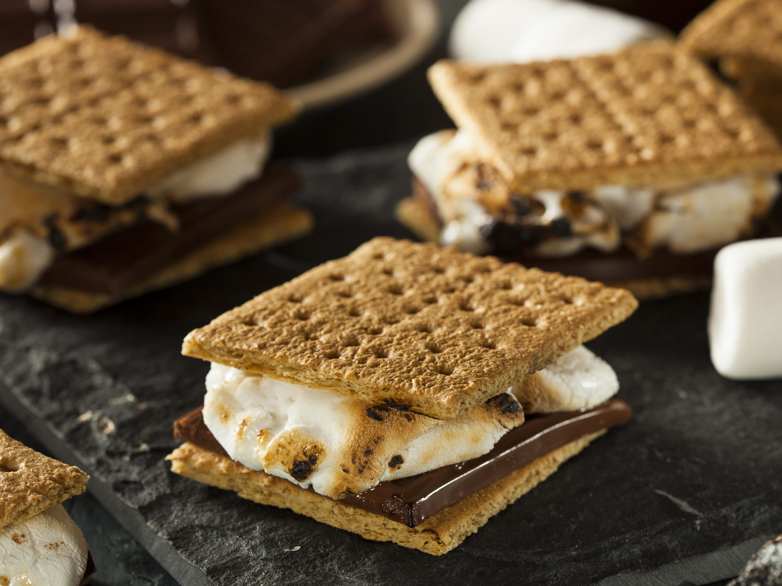 A counter top hosts seven s'mores featuring graham crackers, white marshmallows and thick chocolate slices.  The s'more dessert sandwiches are thick.  Each graham cracker, marshmallow filling and dark chocolate filling rests at a slightly different angle causing every dessert sandwich to look unique.